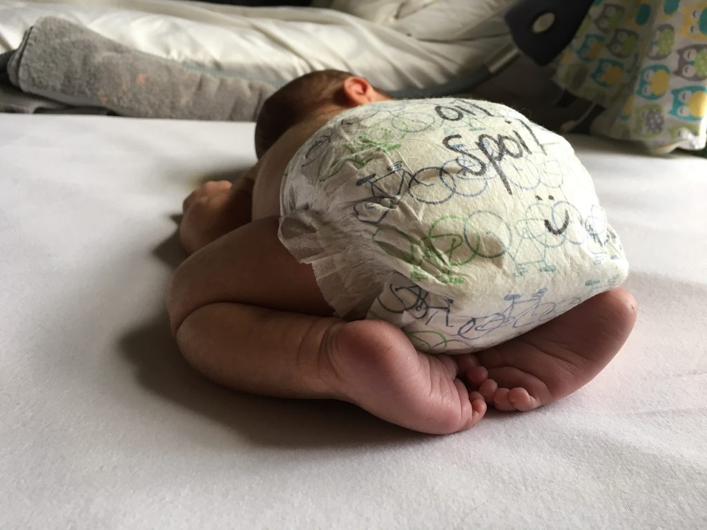 A baby laying on his stomach in nothing but a diaper, facing away from the camera. The baby's feet are tucked up under his bottom, and his diaper has "Oil Spoil" written on it in black marker. 