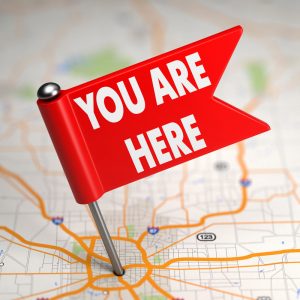 A pin labeled "You Are Here" sticking into an atlas-style road map, illustrating the need to identify your financial starting point prior to charting a course to your financial goals.